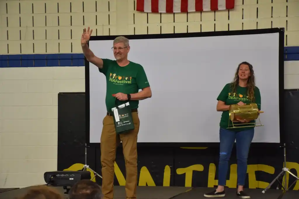 Man in green shirt waves, woman beside him holds a drum.  Both are on stage at an event.