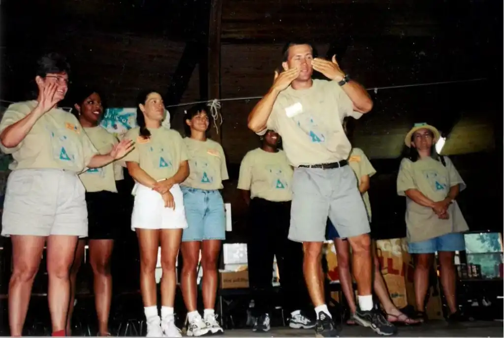 A group of camp staff stand on a stage wearing matching tan shirts. One man in the center is signing to a crowd.
