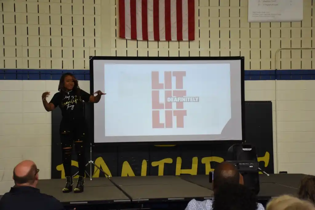 A woman giving a presentation on a stage. The presentation's title is "LIT" repeated three times.  She is gesturing energetically.