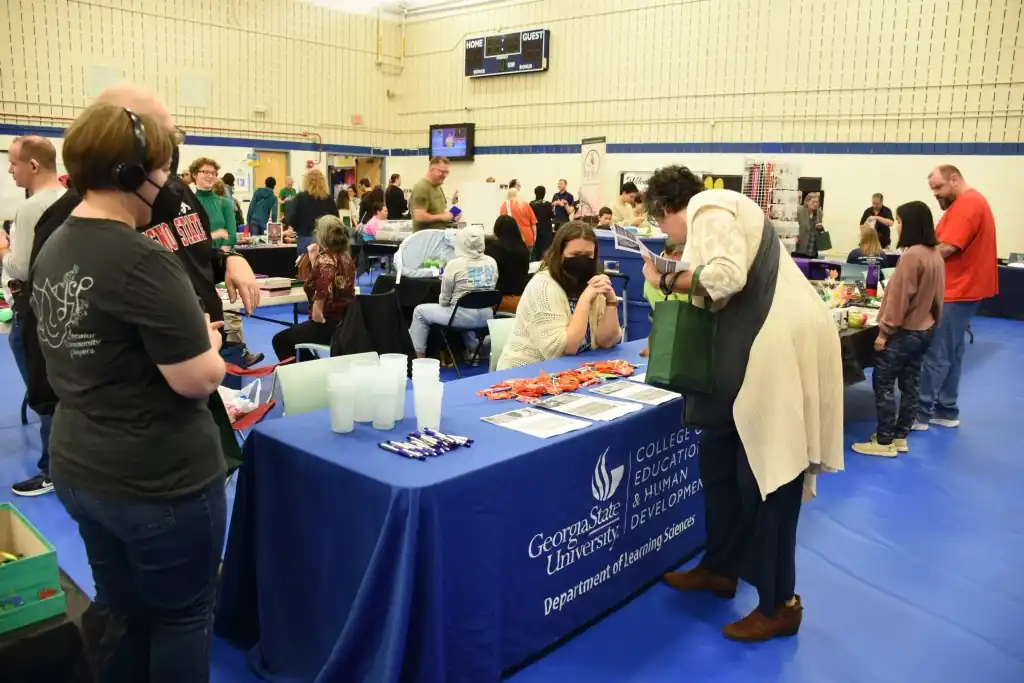A college fair in a gymnasium.  People browse tables with information, some wearing masks.  A table displays the George Mason University logo.