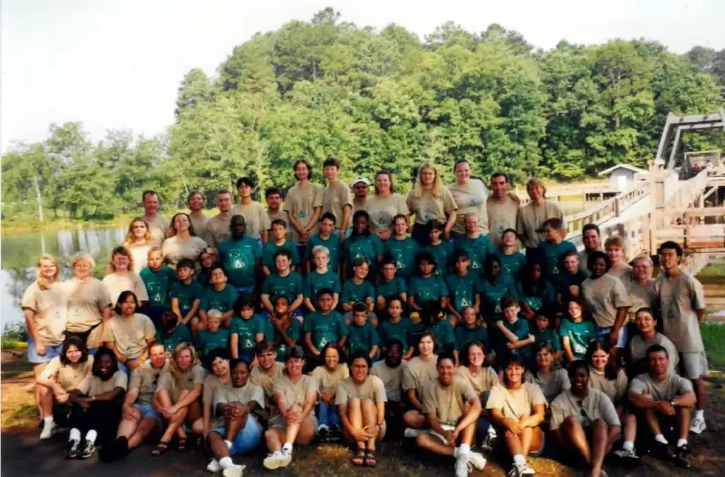 Large group photo of children and adults at Camp Juliena.  They are wearing teal and tan t-shirts.  A lake and woods are in the background.