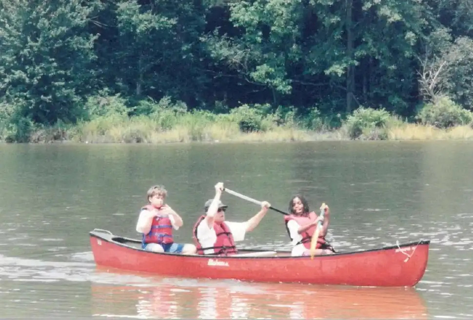 Three people in a red canoe on a calm lake, paddling and enjoying a sunny day. Lush green trees line the shore.