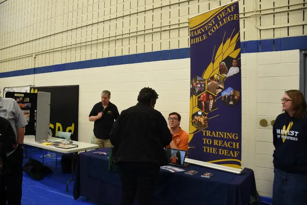 Representatives from Harvest Deaf Bible College talk with prospective students at a college fair.  A banner displays the college's logo and mission: "Training to Reach the Deaf."