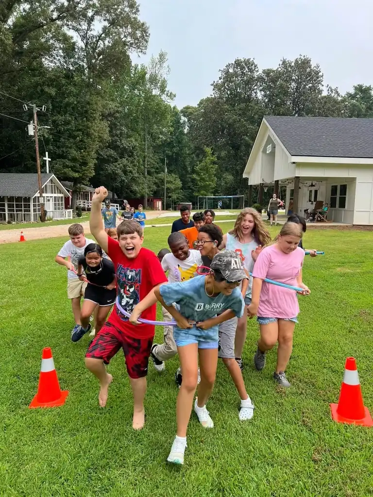 A group of kids playing a game outdoors on a grassy field. They are holding a long ribbon and running. Two orange cones are next to them.