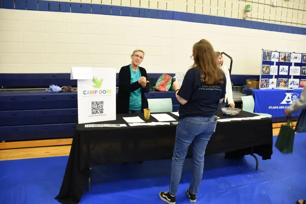 A woman at a table with informational pamphlets talks to a woman at a school event.  A QR code is visible on the table.