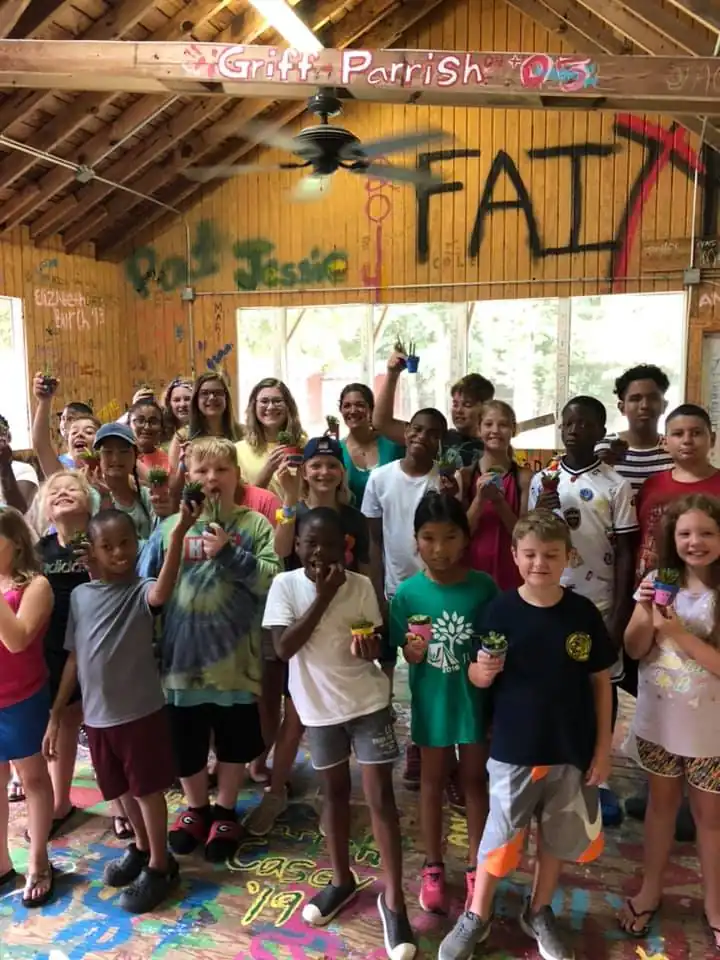 A large group of children and teenagers stand in a room with graffiti-covered walls, holding small potted plants. They are smiling at the camera.