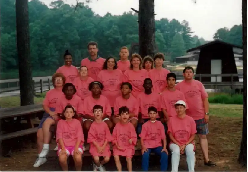 Group photo of campers and staff wearing pink Camp Juliena t-shirts, posing outdoors near picnic tables and trees.