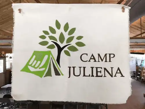 Hanging sign of Camp Juliena logo which shows a tree and a green tent with a white ILY icon on the side of it.