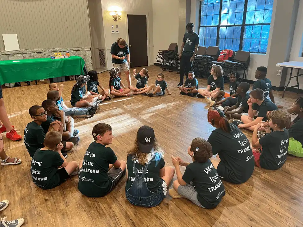 Campers sit in a circle inside a light filled gymnasium. All are wearing matching Camp Juliena shirts.
