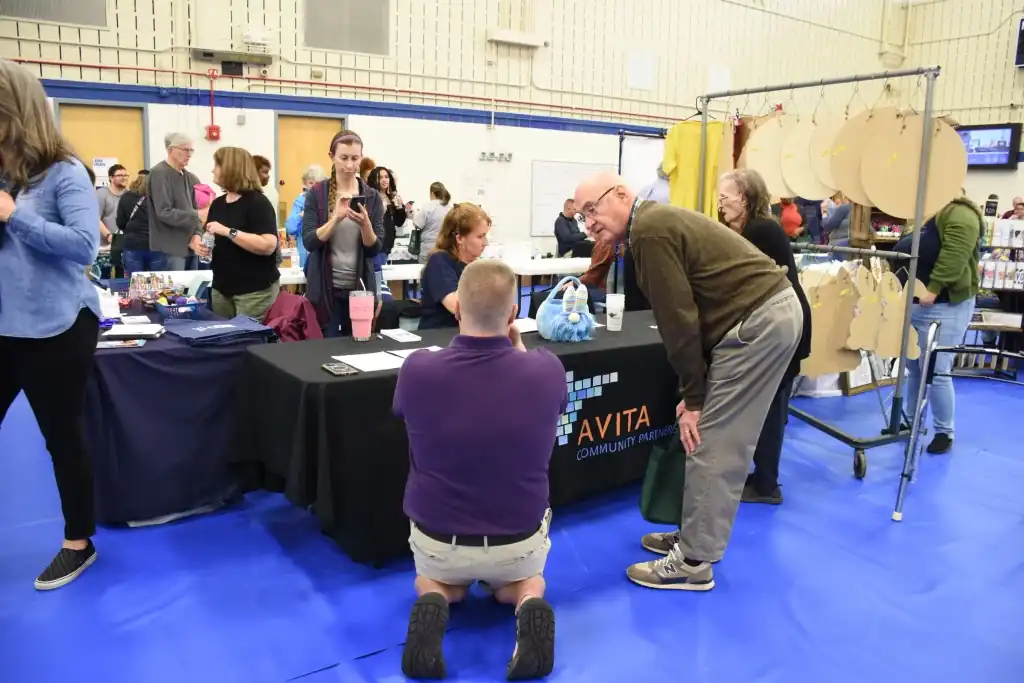 A man kneeling at a table at a crowded event, talking to an older man.  Other people are standing around, looking at displays.