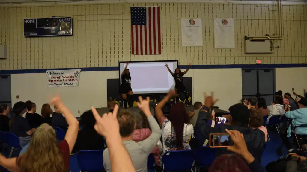 Two performers on stage in front of an audience. The audience is cheering and raising their hands in the ILY sign.  A large screen is behind the performers.