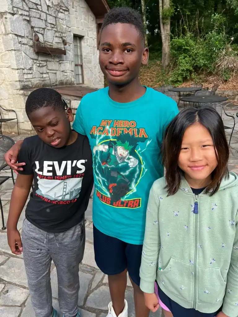 Three campers pose for a photograph, two boys and one girl. A stone wall and trees are in the background.