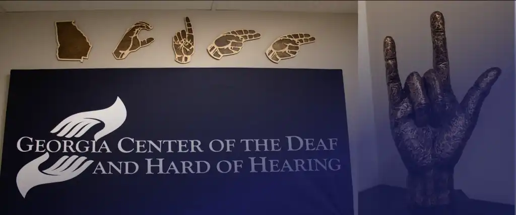 Banner with two images: GCDHH wall decoration, blue sign with GCDHH logo, wood hangings in the shape of the state of GA, and hands spelling out GCDHH. ILY hand statue next to it. Blue overlay.