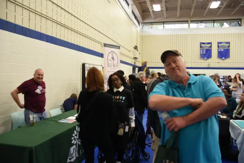 A man in a teal shirt stands with his arms crossed, looking at the camera.  Other people are in the background at what looks like an event or fair.
