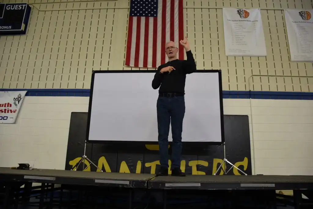 A man giving a presentation using sign language in front of a projector screen in a gymnasium.