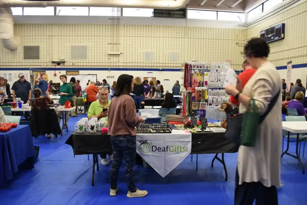 A craft fair in a gymnasium. Several vendors are at tables displaying their goods. A woman is looking at jewelry at the DeafGirls! booth.