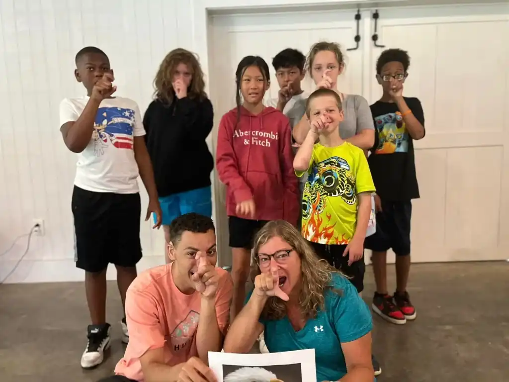 A group of campers and staff pose for a photo, signing EAGLE, as the staff members hold a picture of an eagle.