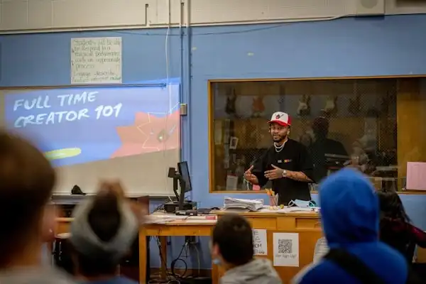 A man stands in front of a classroom wearing a black shirt and red hat. The screen next to him shows a Powerpoint reading "Full Time Creator 101". Students watch him present.