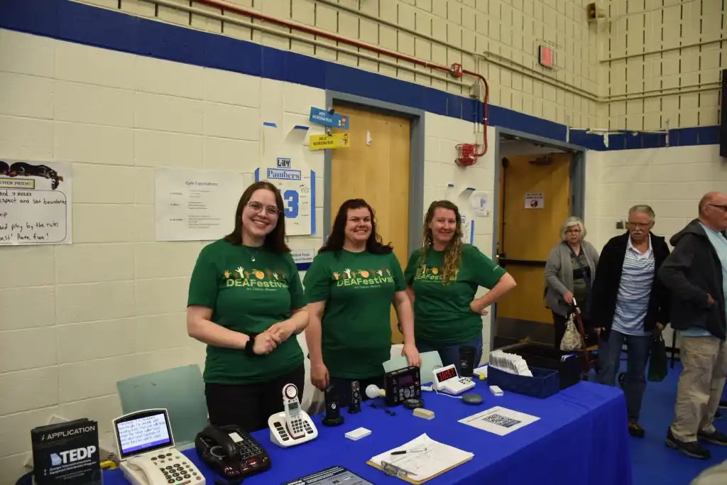 Three women smile at a table with assistive listening devices. They wear green DEAFestival shirts with a logo.  People are in the background.