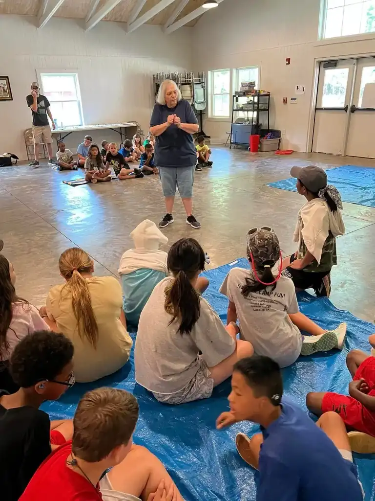 A woman addresses a group of campers sitting on a blue tarp inside a building with several windows.