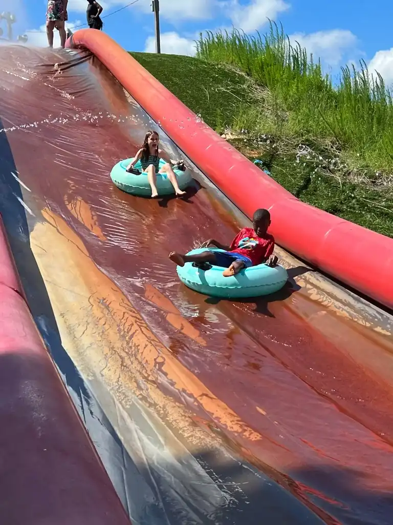 Two children, a girl and a boy, ride inner tubes down a large water slide. The slide is multicolored, with a red inflatable border next to it.