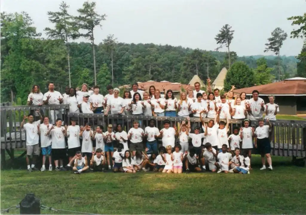 Large group photo of campers and staff at summer camp.  Everyone is wearing white Camp Juliena shirts.  They are posing on a wooden deck in front of camp buildings.