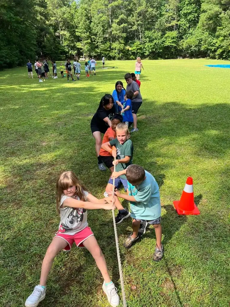 Campers participate in a tug-of-war competition on a grassy field.  Two teams pull on a rope, with a bright orange cone nearby.