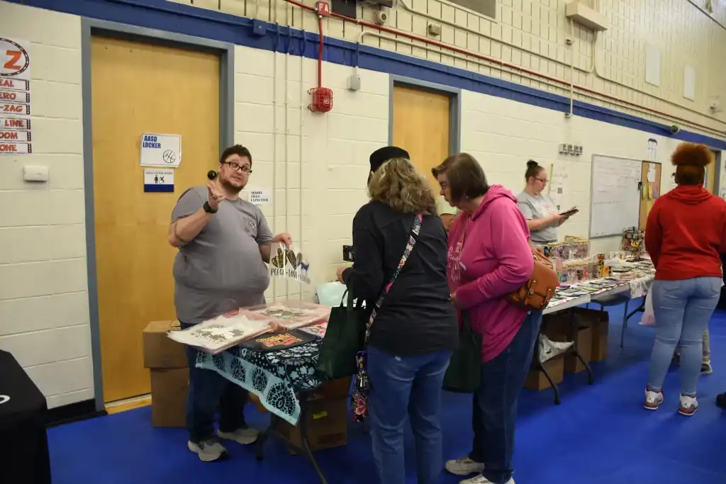 A vendor at a comic book convention talks to customers at his table, showing off merchandise.