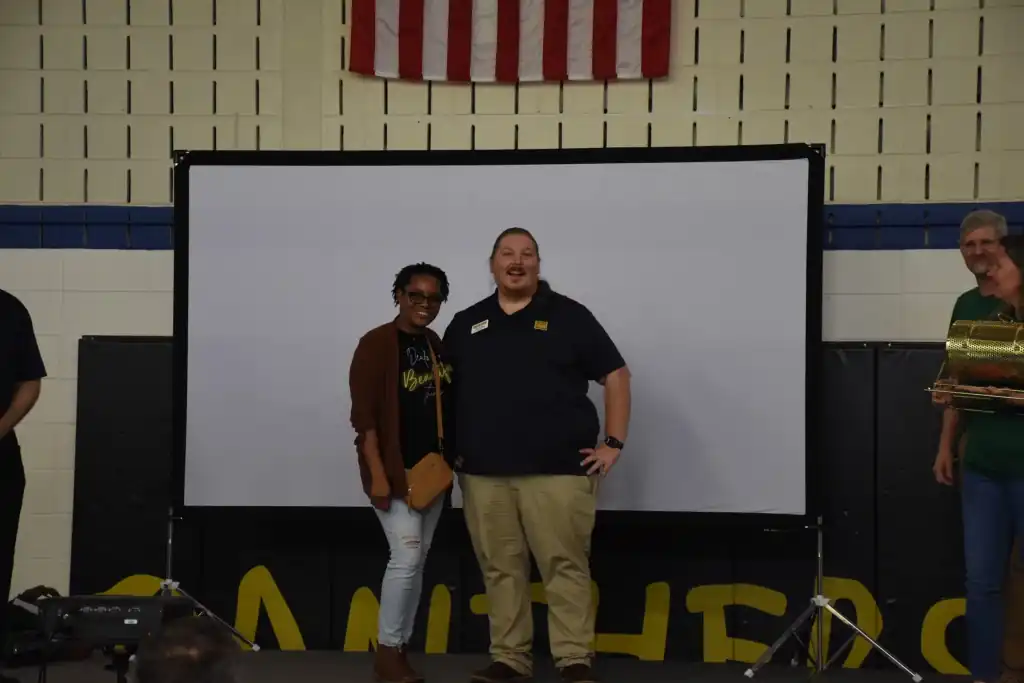 Two people stand in front of a large screen, smiling at the camera. A man and a woman are posing for a photo.