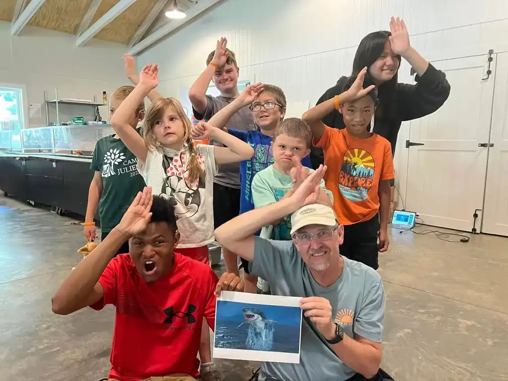 A group of campers and staff pose for a photo with a picture of a shark.  They are holding their hands up to their foreheads to sign SHARK.