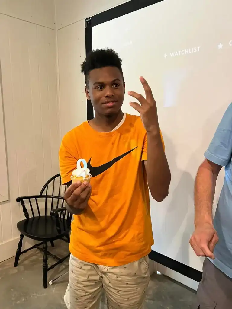 A young man in an orange shirt holds a cupcake. He is standing in front of a projection screen.