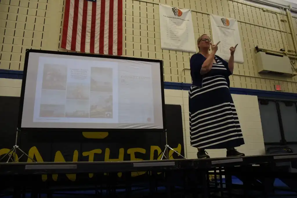A woman in a striped dress gives a presentation, using sign language, in front of a projection screen showing images.