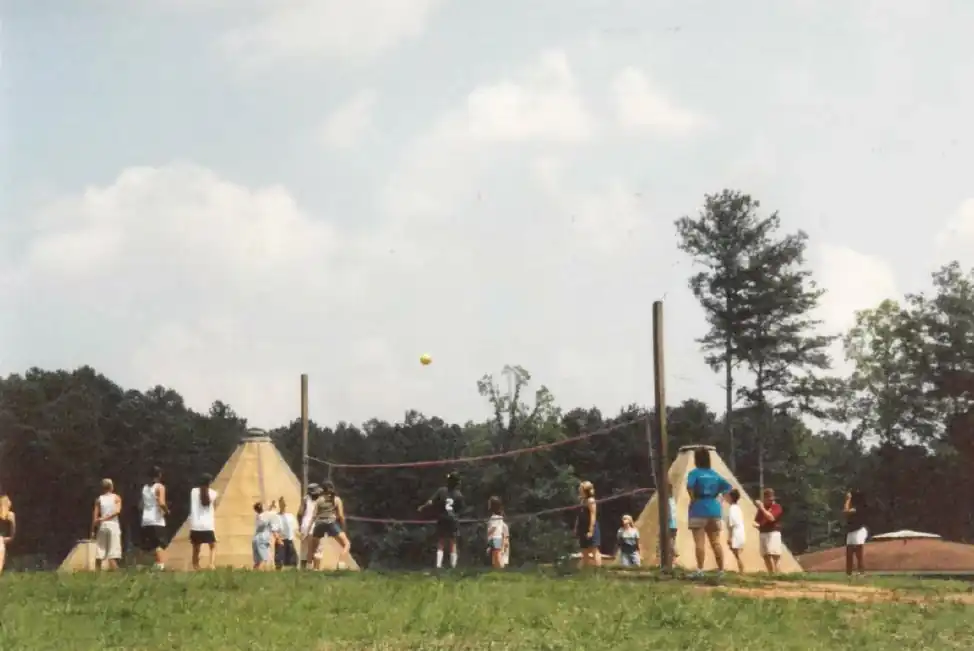 Campers playing volleyball outdoors between two teepees.  A sunny day with trees in the background.