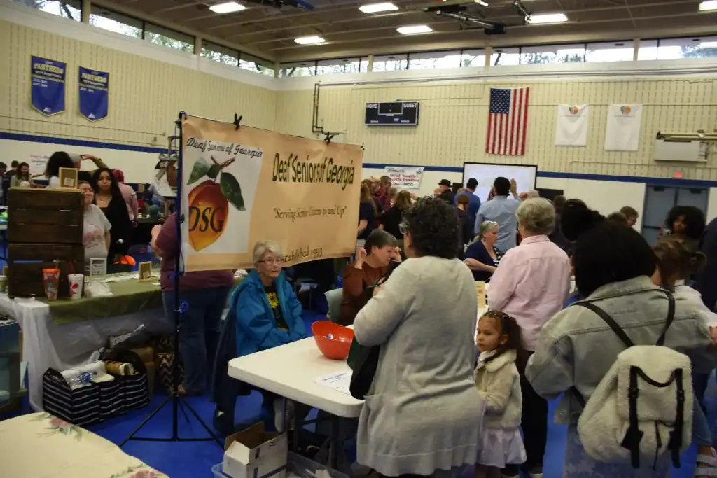 A bustling indoor market scene, with numerous people browsing various vendor tables.  A banner for "Del Seymour Georgia" is prominently displayed.