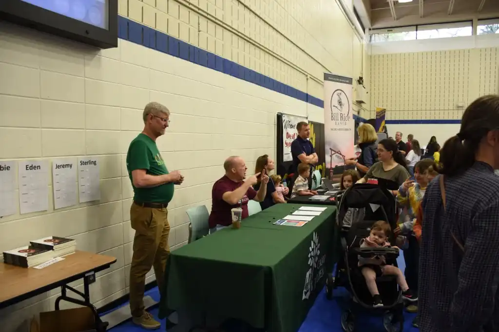 Authors at a book signing, sitting behind a table with books and promotional materials. A crowd of people are waiting in line.