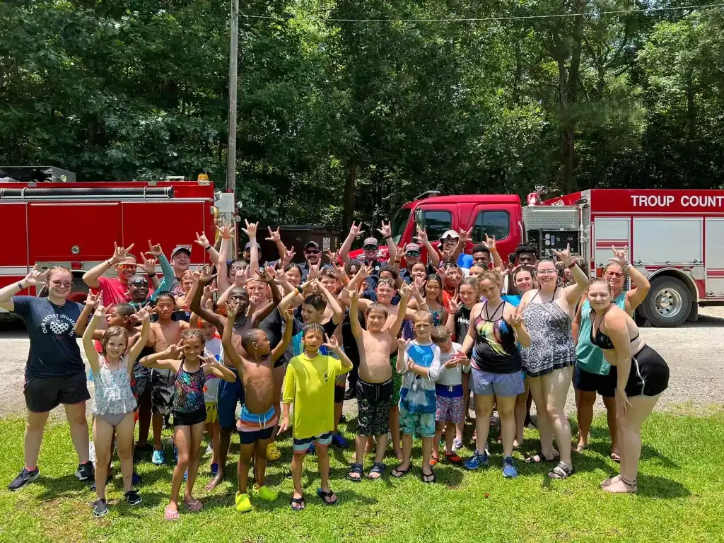 Large group of campers of diverse ages and ethnicities stand in front of a firetruck, making the ILY sign. Many are wearing swimsuits